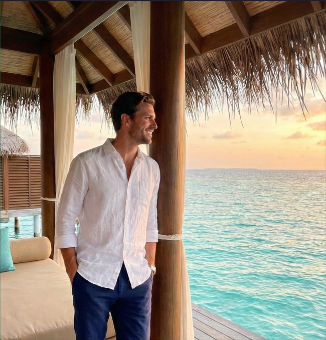 Un homme souriant profite d'un coucher de soleil depuis la terrasse d'un bungalow sur pilotis face à une mer turquoise.